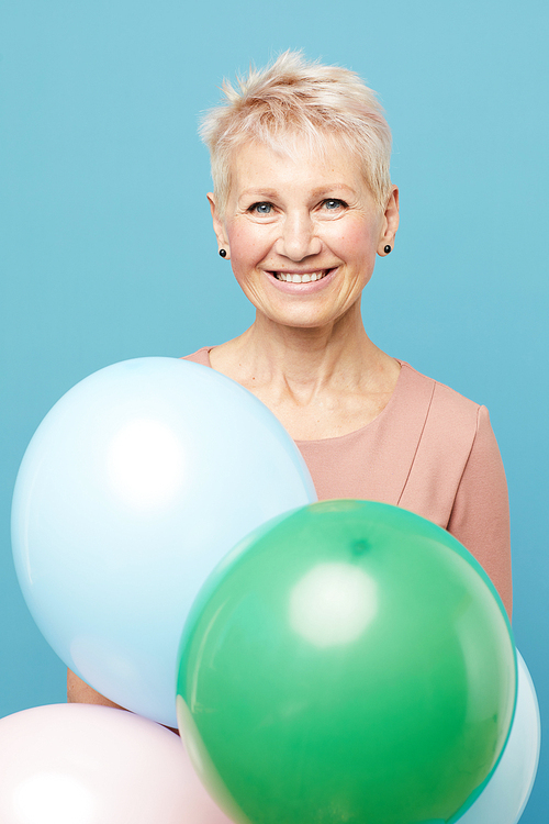 Portrait of jolly beautiful mature lady with short hair standing against blue background and holding colorful balloons
