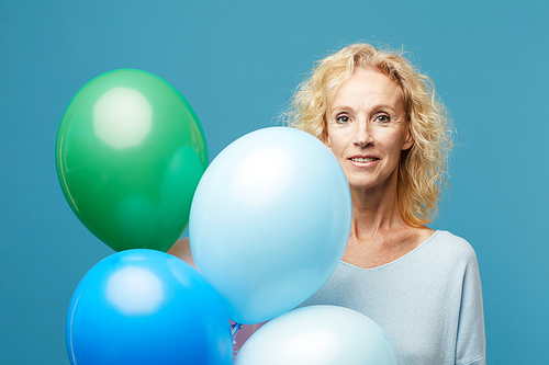 Smiling beautiful mature lady with curly hair holding colorful helium balloons and looking t camera