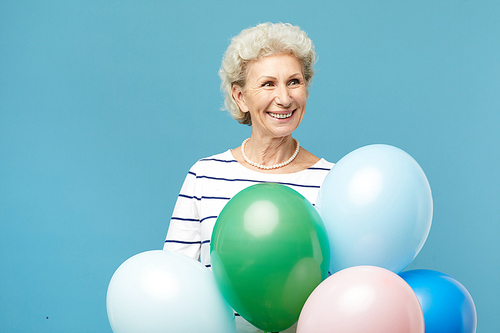 Happy excited elderly lady with curly hair standing against blue wall and holding bouquet of balloons