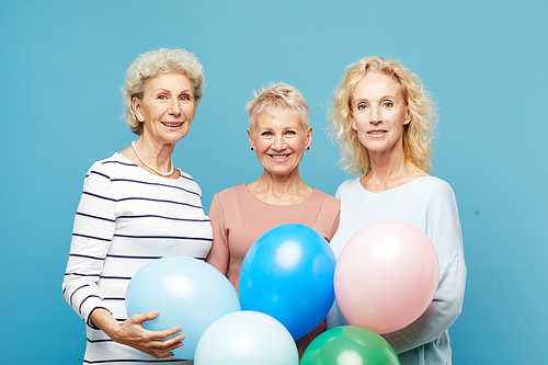 Portrait of smiling mature ladies in casual outfits standing together against blue wall and holding balloons