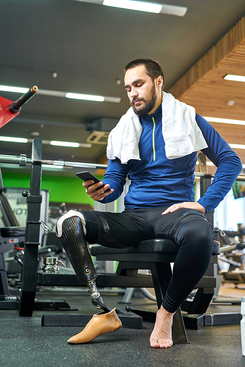 Young athlete with amputee leg sitting in gym and communicating online  using his smartphone