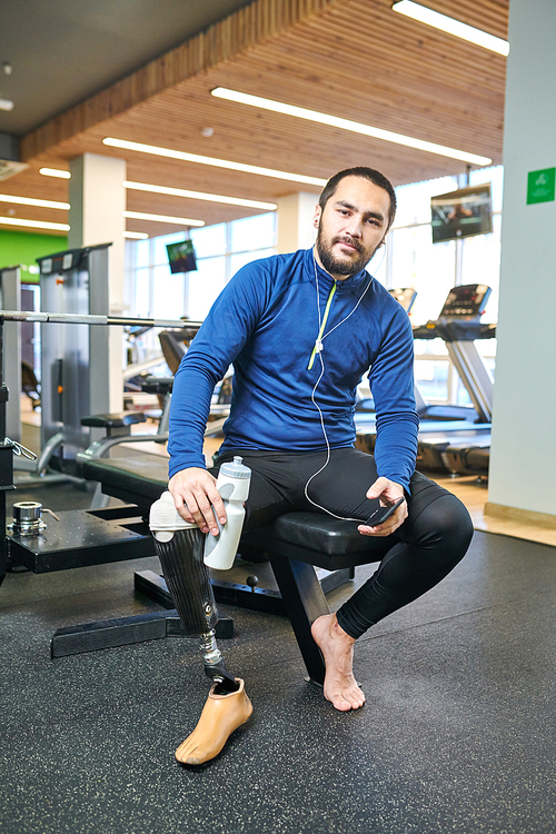 Portrait of bearded disabled athlete listening to music on smartphone and drinking water in fitness club