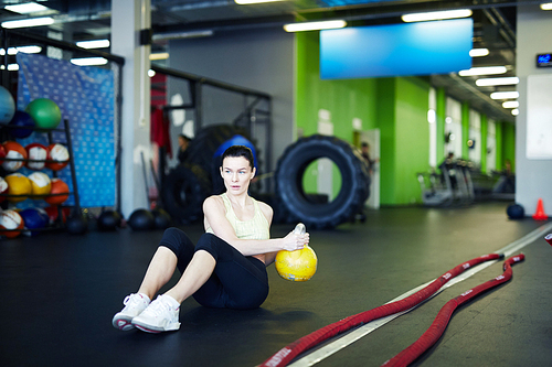 Young sportswoman with kettlebell sitting on the floor of fitness center and doing side turns