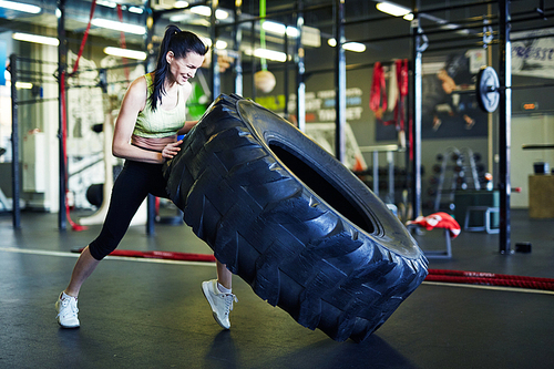 Young strong active woman in sportswear lifting big heavy tyre while training in leisure center