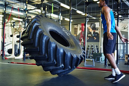 Young athlete in sportswear standing in gym in front of big heavy tyre falling down during workout