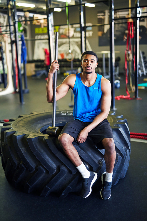 Young confident cross training instructor with hammer sitting on big heavy tyre in front of camera