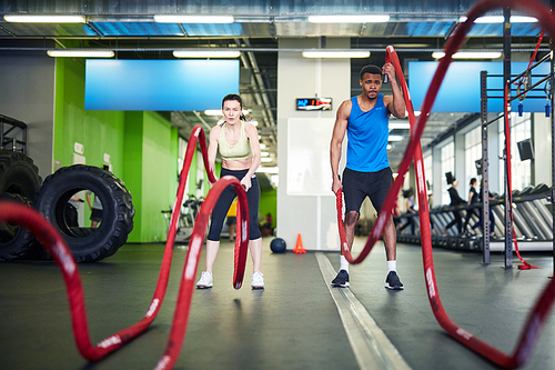 Active young man and woman working out with heavy ropes in gym during cross training