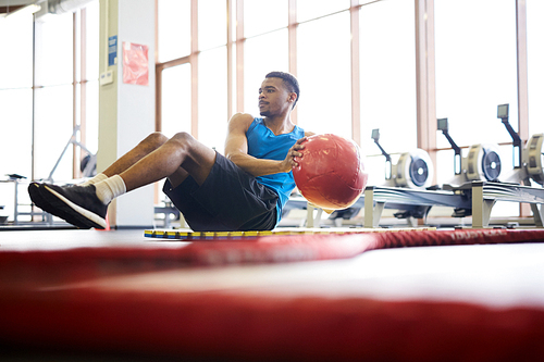Young athlete holding sports ball and turning aside while sitting on mat and exercising in fitness center