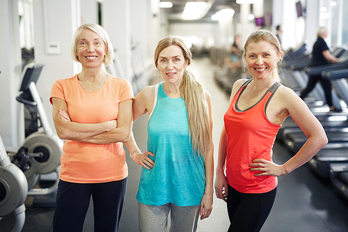 Three happy mature sportswomen in activewear standing in front of camera in contemporary fitness center