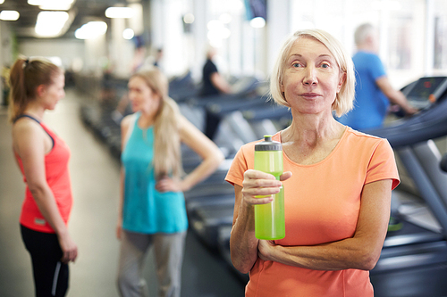 Mature blonde woman in sportswear holding plastic bottle with water while having break after workout in gym