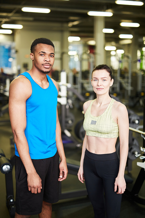 Two young professional sports trainers in activewear standing in front of camera in gym with fitness equipment on background