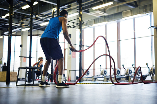 African-american sportsman in activewear holding two ropes while exercising in fitness center