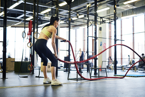 Young active woman with bent knees standing on the floor and exercising with cross training ropes in gym