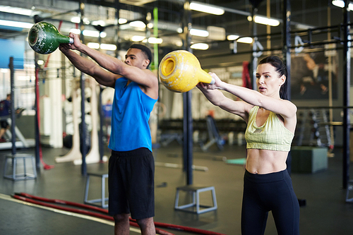 Two young athletes in activewear lifting heavy kettlebells while working out in fitness center