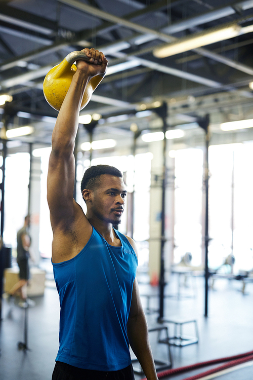 Young athlete in activewear stretching upwards right arm with heavy kettlebell while exercising in gym