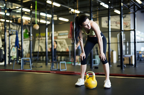 Young active woman bending and looking aside while having short break after difficult exercise in gym