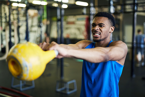 Young strong athlete making effort while exercising with heavy kettlebell in fitness center or gym
