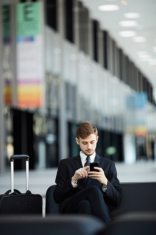 Portrait of young businessman using smartphone while relaxing in VIP lounge of airport waiting room, copy space