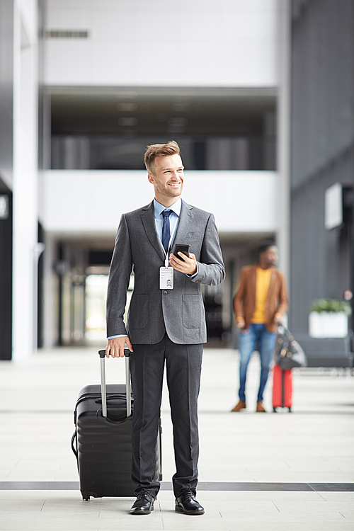 Cheerful handsome young businessman with badge hanging on neck standing with luggage and using phone, he arriving in airport