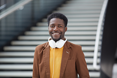 Portrait of cheerful hipster black guy with beard wearing wired headphones on neck standing against stairs