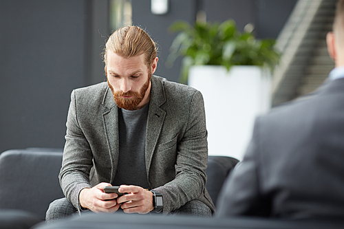 Serious young bearded man in gray jacket sitting in lobby and using internet app on smartphone