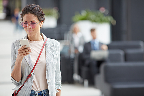 Content attractive stylish girl in sunglasses wearing stripped jacket and red bag checking phone on move