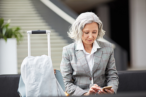 Smiling gray-haired Asian lady in checkered jacket sitting in airport and checking messenger on phone