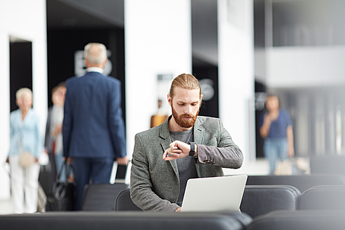 Serious young bearded entrepreneur sitting with laptop in airport and looking at wristwatch while checking time till boarding