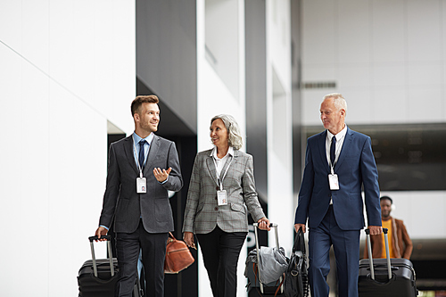 Group of confident company managers in suits walking with luggage in airport and discussing business trip