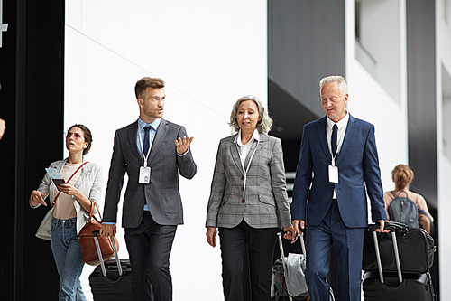 Group of multi-ethnic business people in suits moving along airport with suitcases and listening to young colleague
