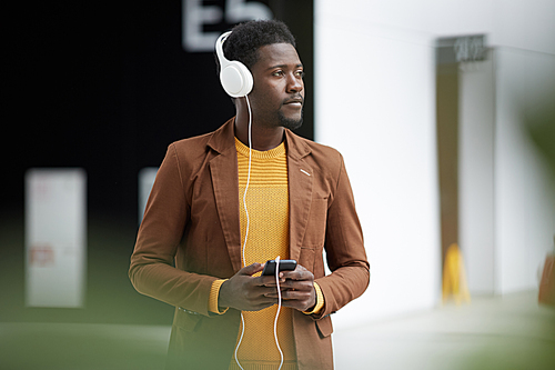 Pensive dreamy young black man in casual jacket standing in lobby and listening to audiobook in headphones