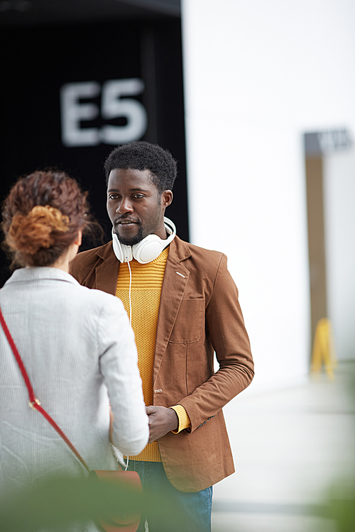 Content handsome Black man with headphones on neck standing in airport and talking to girl