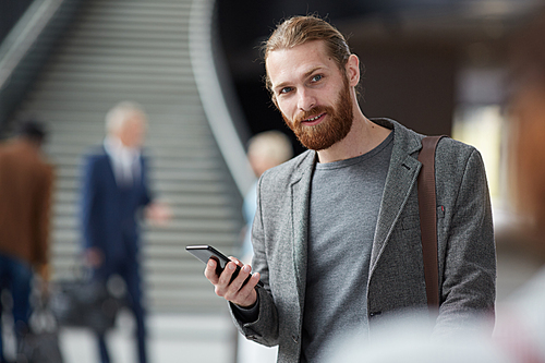 Portrait of positive red-bearded guy in gray jacket standing in lobby and using gadget