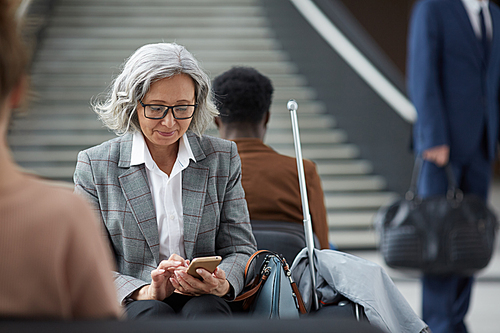 Content elderly Asian woman with gray hair wearing eyeglasses sitting in waiting area of airport and using mobile app on gadget