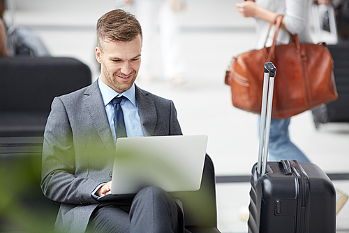 Smiling young marketer with stubble wearing gray suit sitting in airport and working with online data on portable computer