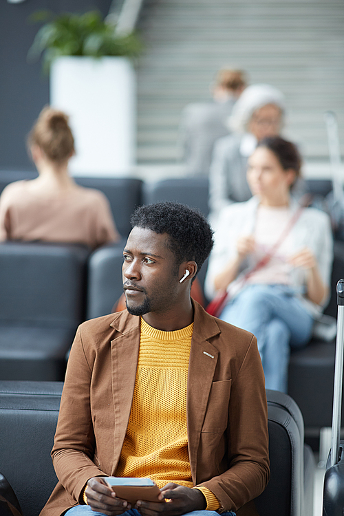 Serious pensive young Afro-American man in brown jacket sitting on sofa in airport and listening to audio in wireless earphones waiting for boarding