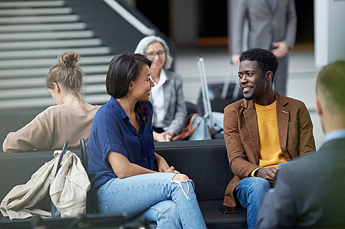Positive handsome young Afro-American man with beard sitting on sofa and talking to lady in airport area