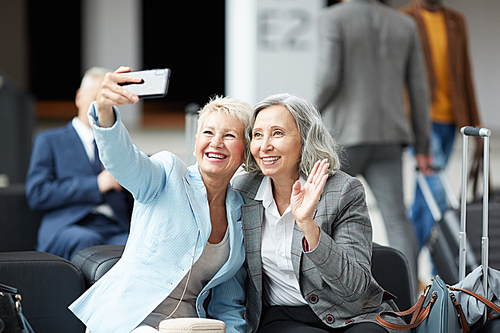 Cheerful mature multi-ethnic ladies sitting on sofa in airport and photographing together on smartphone