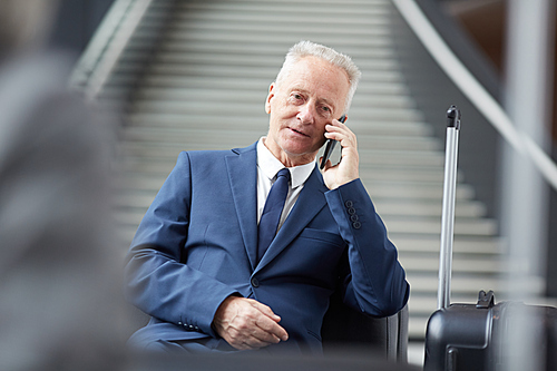 Serious handsome mature businessman in formal suit sitting on sofa in airport and communicating on cellphone