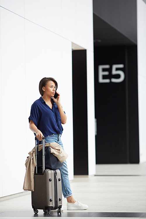 Pensive young woman in casual outfit standing with wheeled suitcase in airport and communicating on phone