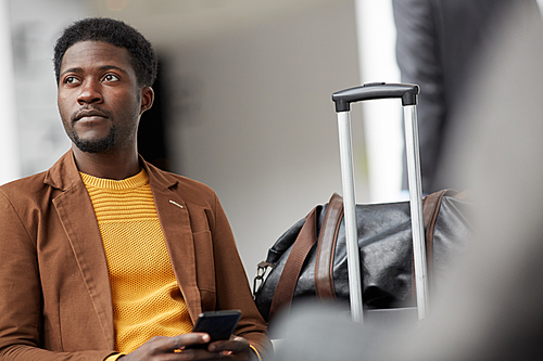 Serious handsome young Black man with beard sitting with luggage in airport and waiting for his airplane