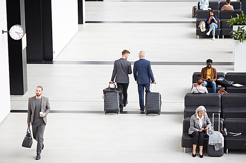 Above view of modern businessmen in formalwear carrying wheeled suitcases while crossing airport waiting area