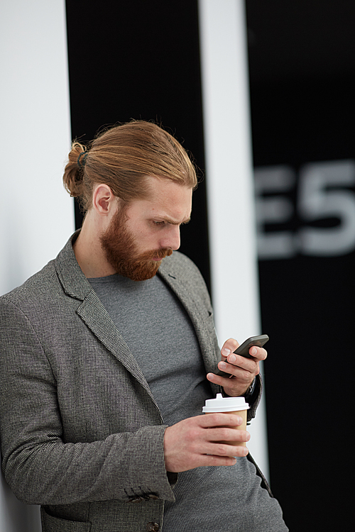 Concentrated red-bearded man in jacket leaning on wall and reading sms on smartphone while drinking coffee in lobby