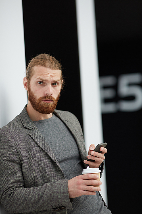 Portrait of serious confused handsome young man with beard using smartphone and drinking coffee