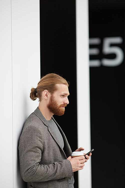 Content hipster bearded man in gray jacket standing at wall in lobby and browsing internet on modern phone