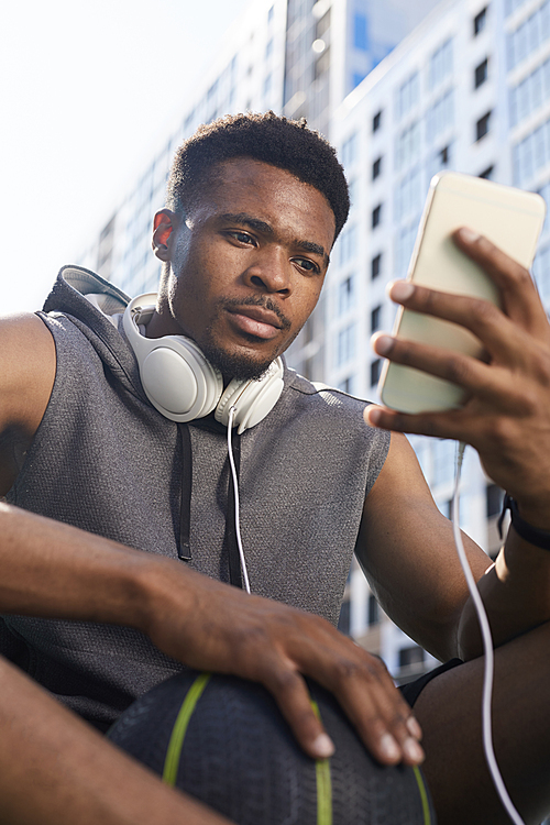 Low angle portrait of contemporary African-American man using smartphone while sitting in basketball court, copy space