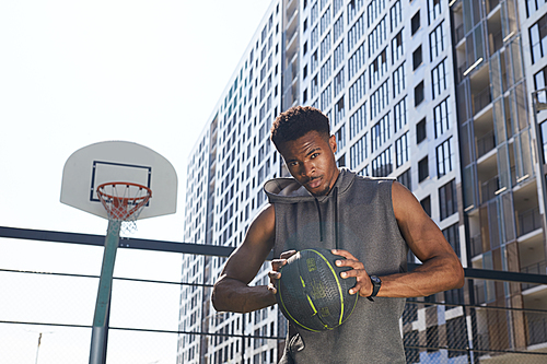 Waist up portrait of handsome African-American man holding basketball ball and  confidently while standing in outdoor court, copy space
