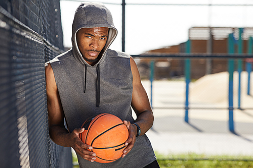 Waist up portrait of contemporary African-American man holding basketball ball  while posing in sports court outdoors, copy space