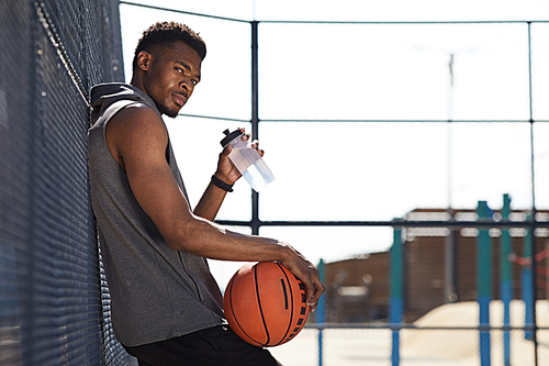 Side view portrait of handsome African-American man holding basketball ball  while drinking water in sports court outdoors, copy space