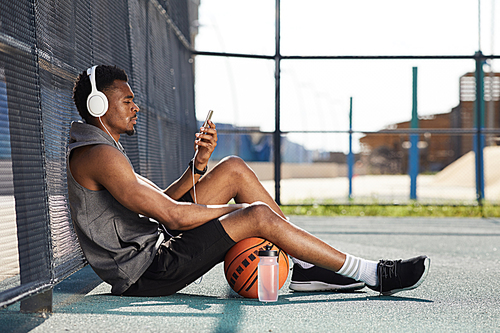 Side view portrait of contemporary African-American man wearing headphones and using smartphone while sitting in basketball court outdoors, copy space